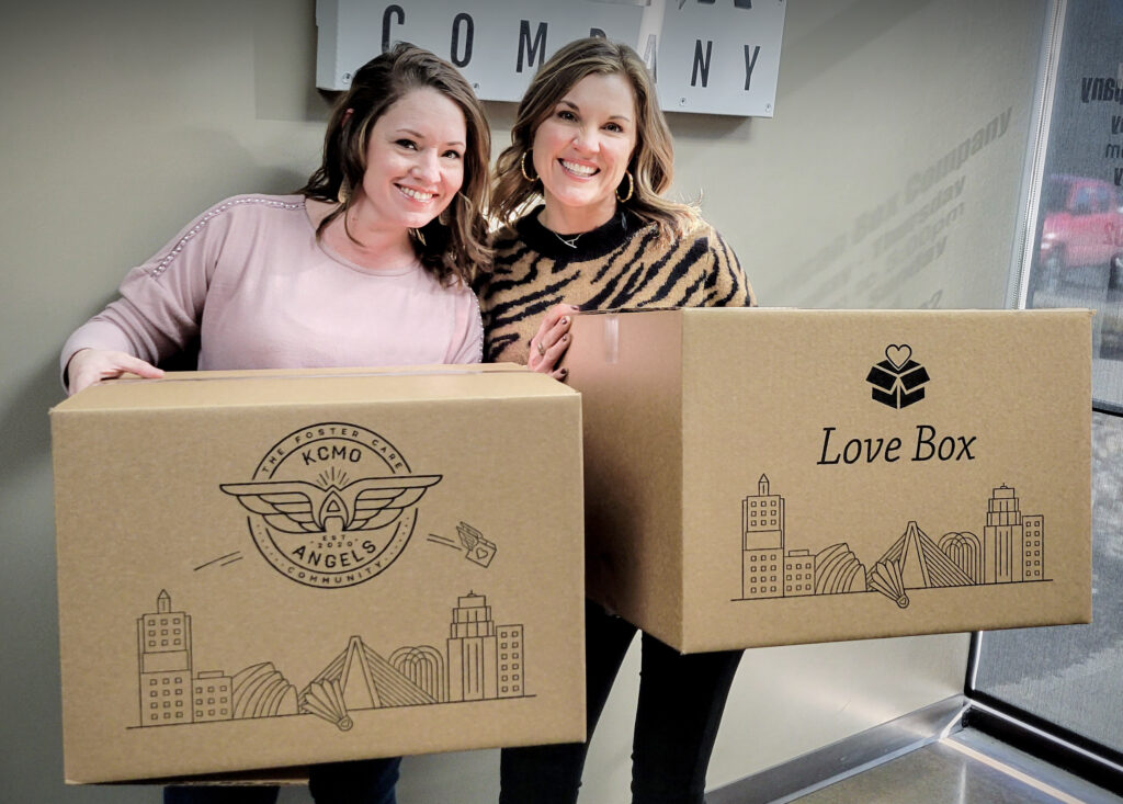 Two women holding corrugated boxes with print showing KCMO Angels logo and KC skyline print