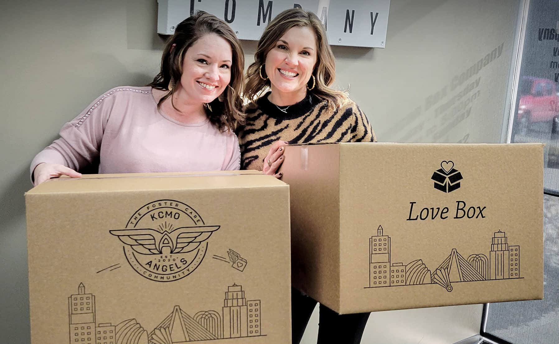 Two women holding corrugated boxes with print showing KCMO Angels logo and KC skyline print